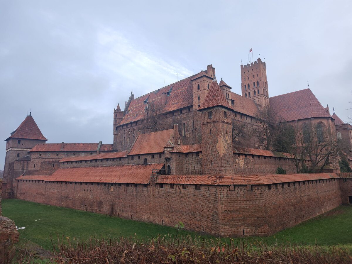 The red brick walls and towers of Malbork Castle across a green moat