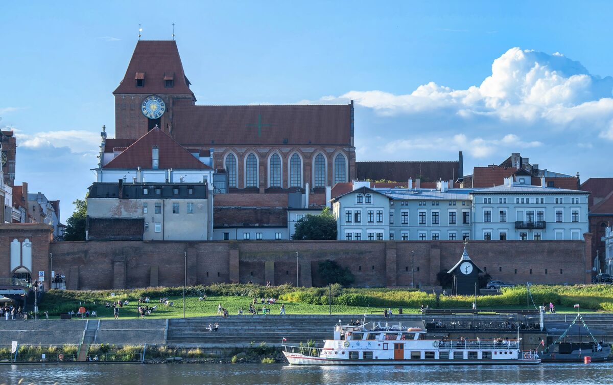 Toruń's Gothic cathedral and old town walls seen from across the Vistula, with a riverboat in front