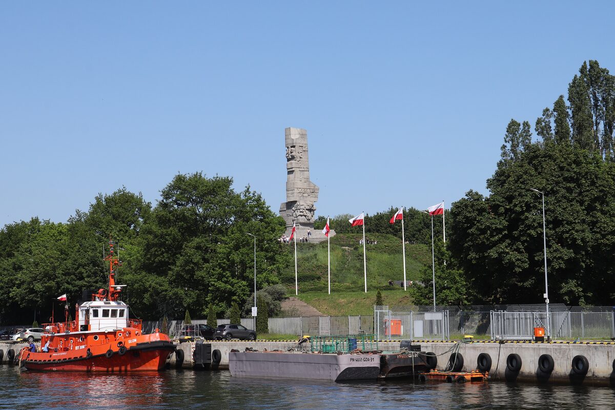 The Westerplatte Monument on its green mound above the harbor channel, with a red tugboat moored in front
