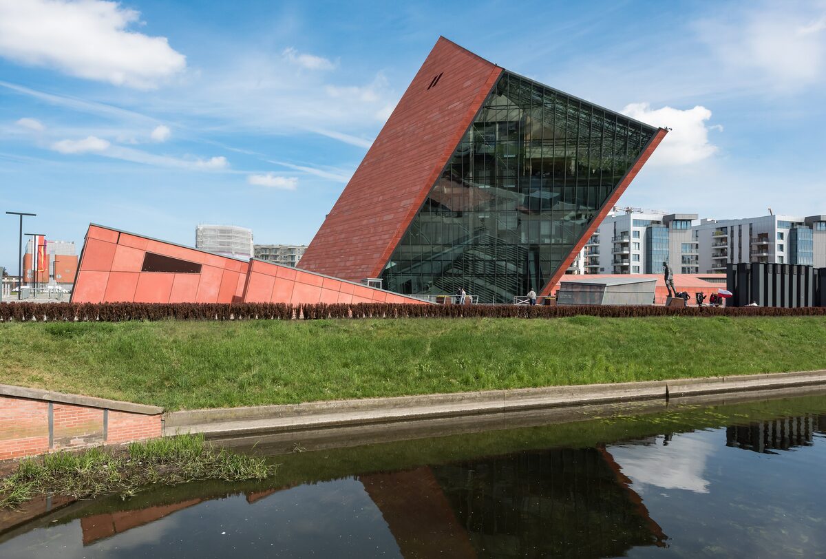 The leaning red tower and glass facade of the Museum of the Second World War, reflected in a canal
