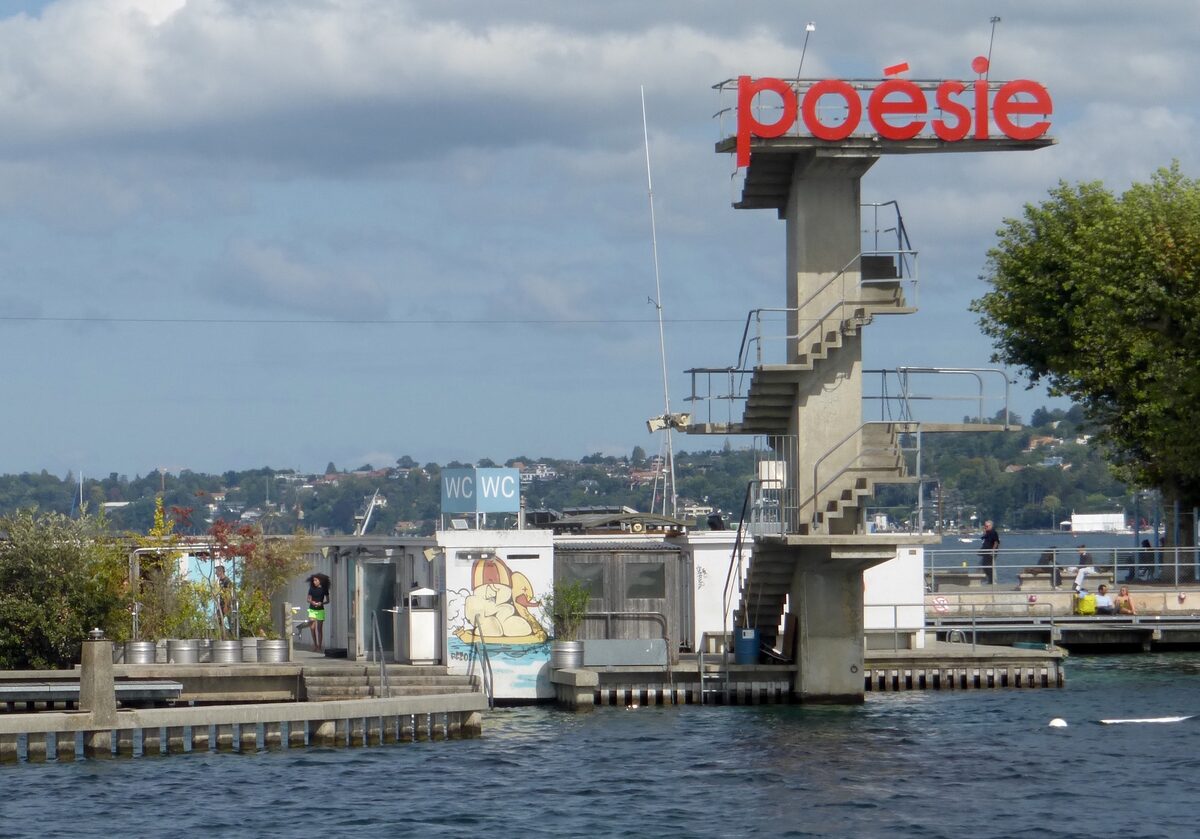 The concrete diving tower of the Bains des Pâquis with its red poésie sign, bathing cabins and a pontoon on the lake