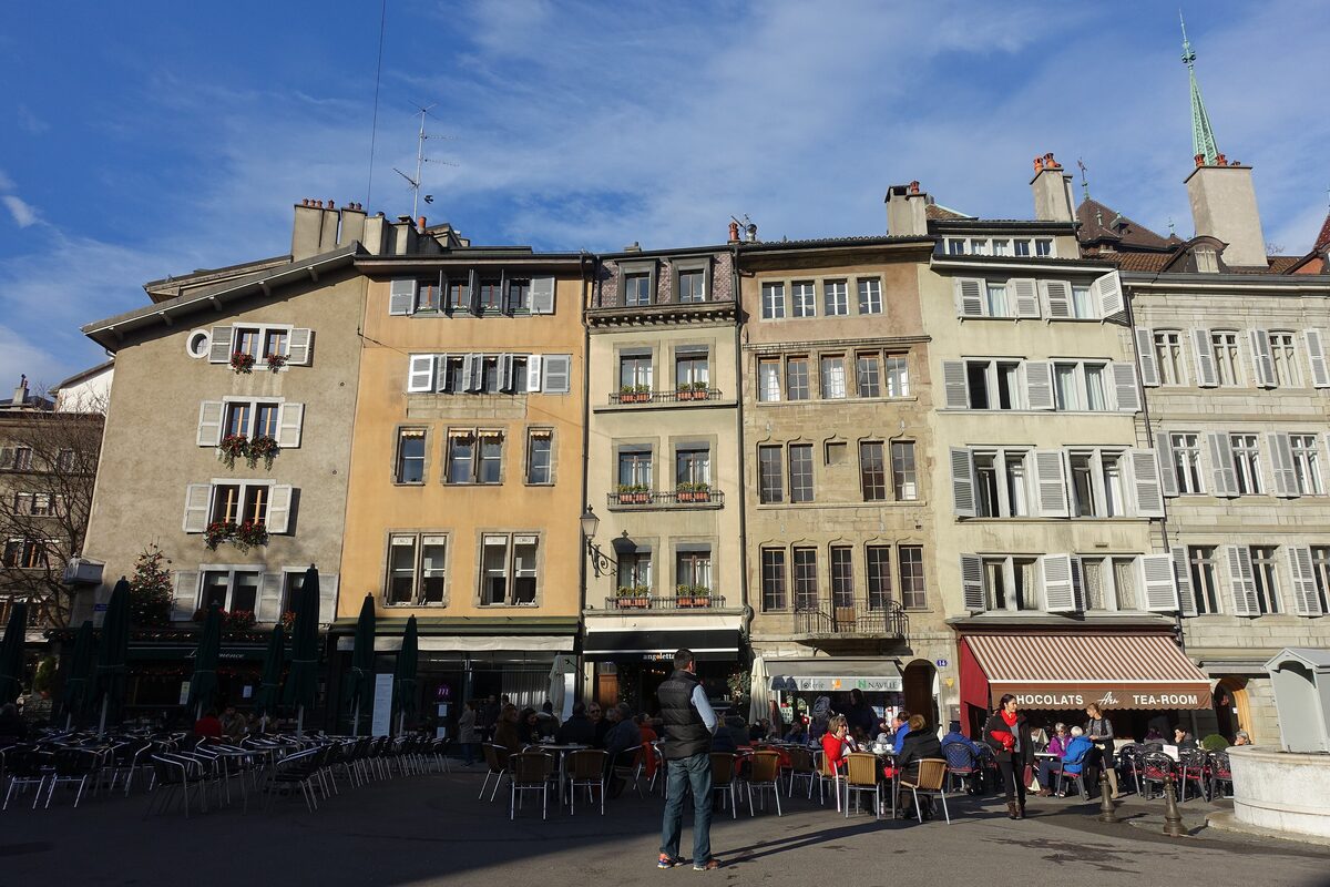 Tall old townhouses in warm stone colors around Place du Bourg-de-Four, with café terraces below and the cathedral spire behind