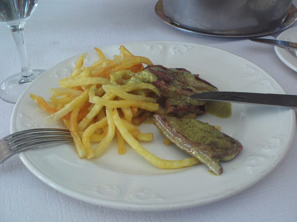 Sliced entrecôte covered in green herb butter sauce with a pile of thin fries on a white plate