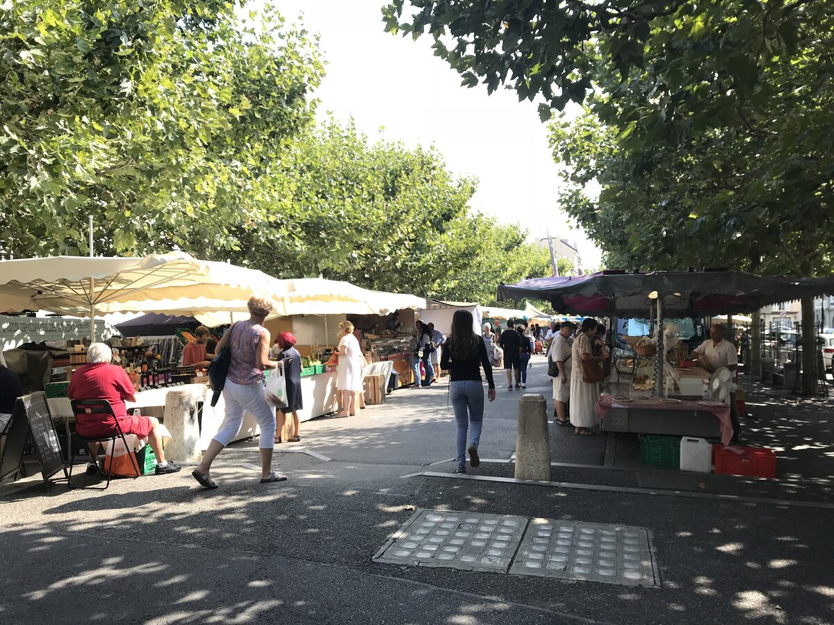 Market stalls and shoppers under the trees on a sunny square in Carouge