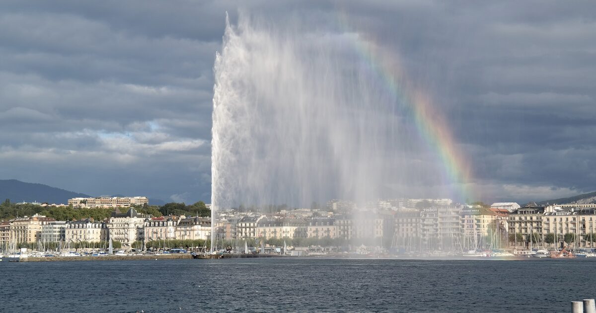 The Jet d'Eau rising from Lake Geneva under a grey sky, with a rainbow beside it and the lakefront buildings behind