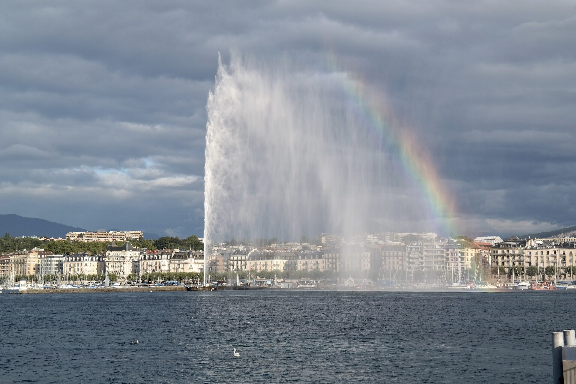 The Jet d'Eau rising from Lake Geneva under a grey sky, with a rainbow beside it and the lakefront buildings behind