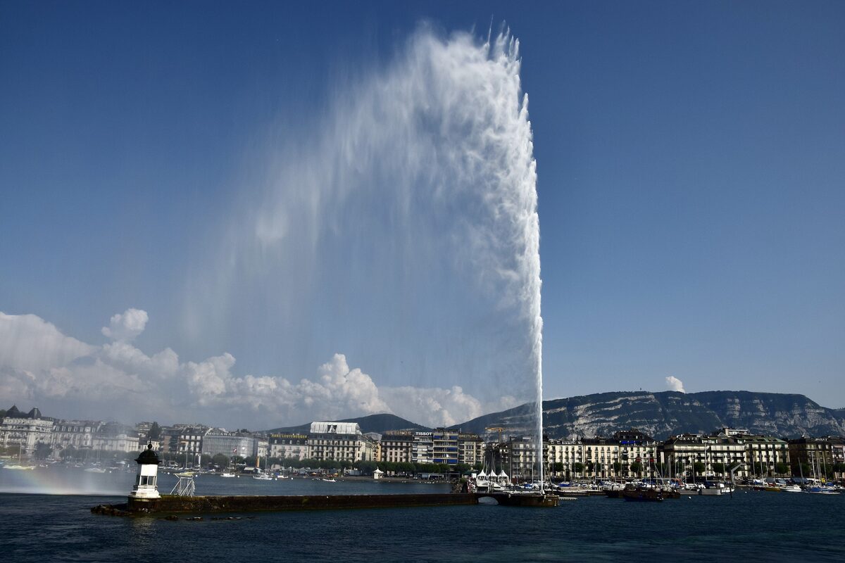 The Jet d'Eau rising high against a blue sky, seen from the water with the small jetty lighthouse and the Geneva waterfront below