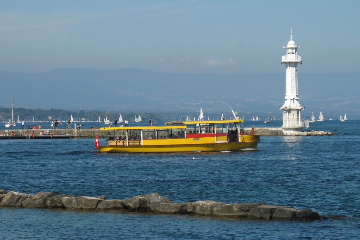 A yellow Mouette shuttle boat crossing the water in front of the white Pâquis lighthouse, with sailboats and mountains behind