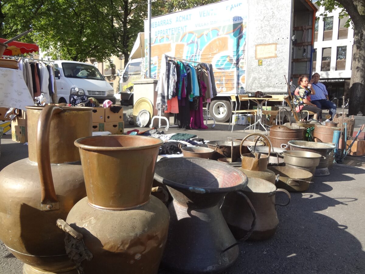 Copper jugs and pots on a stall at the Plainpalais flea market, with clothes racks and a van behind