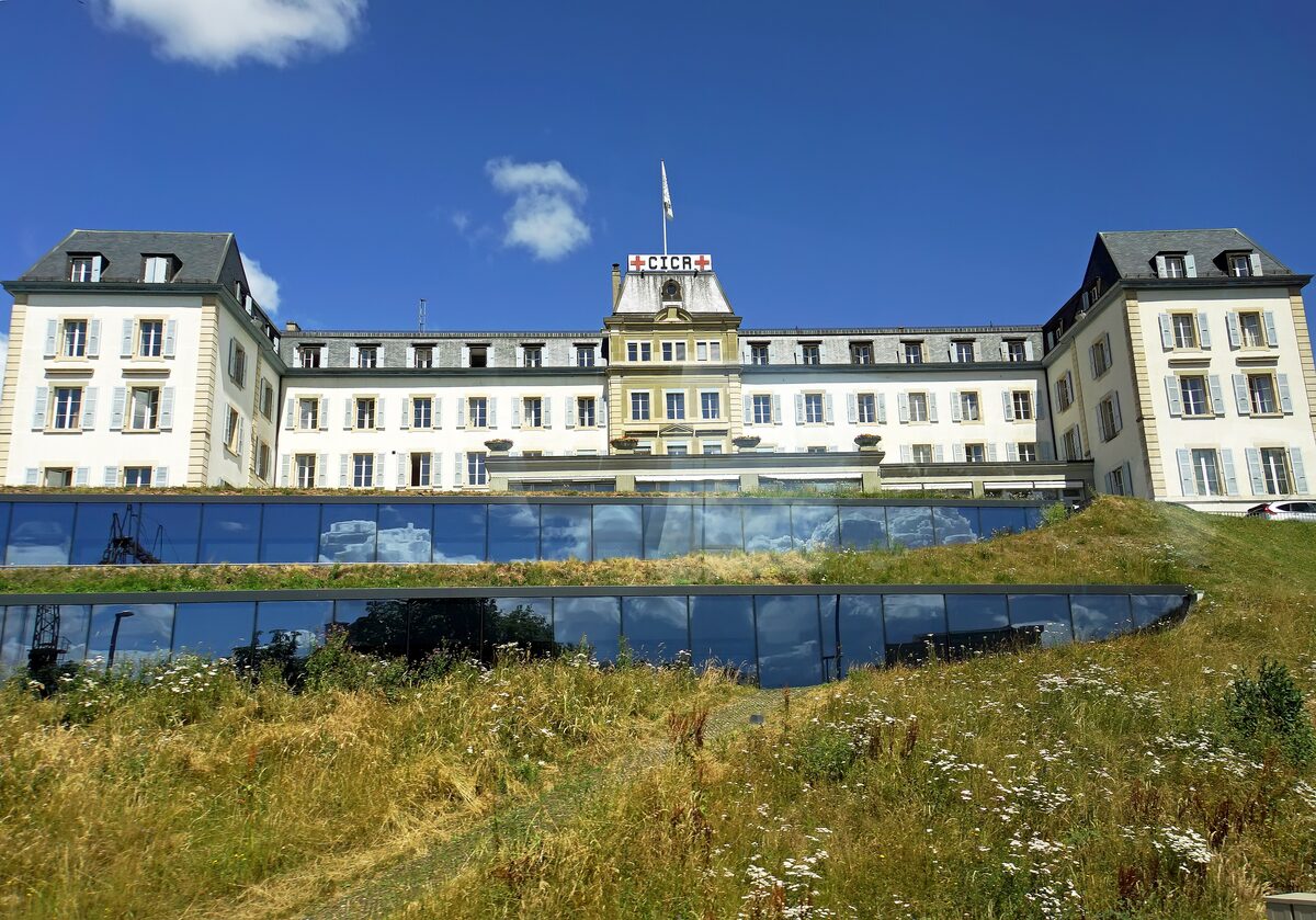 The cream-colored museum building with its red cross on the roof above a grass-covered glass base, under a blue sky