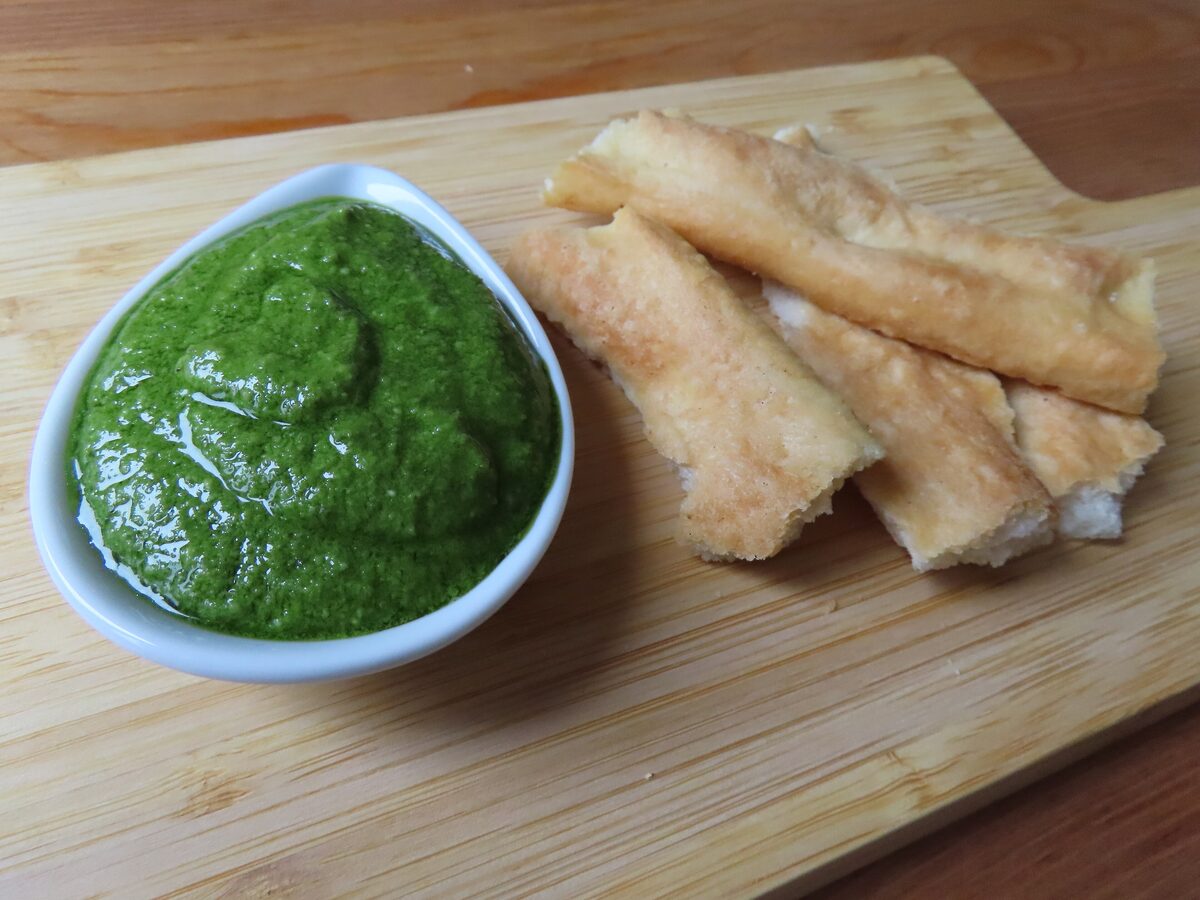 A bowl of bright green pesto beside strips of focaccia on a board