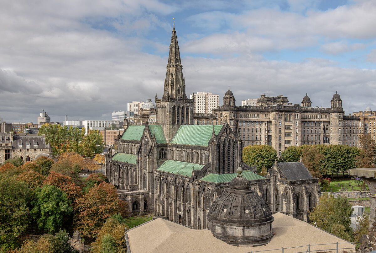 Glasgow Cathedral, a dark stone Gothic church with a spire and green copper roofs, seen from above with autumn trees around it