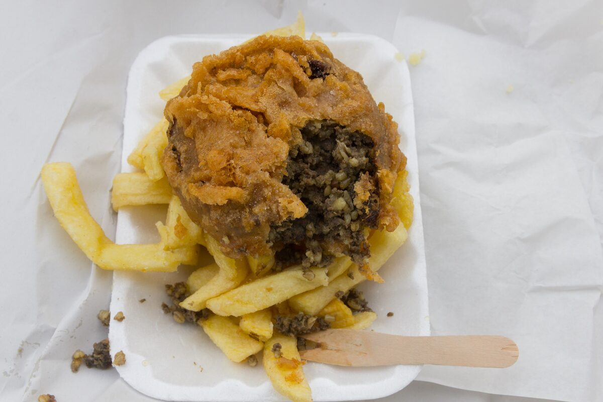 A battered haggis supper with chips on a takeaway tray, cut open to show the dark peppery filling