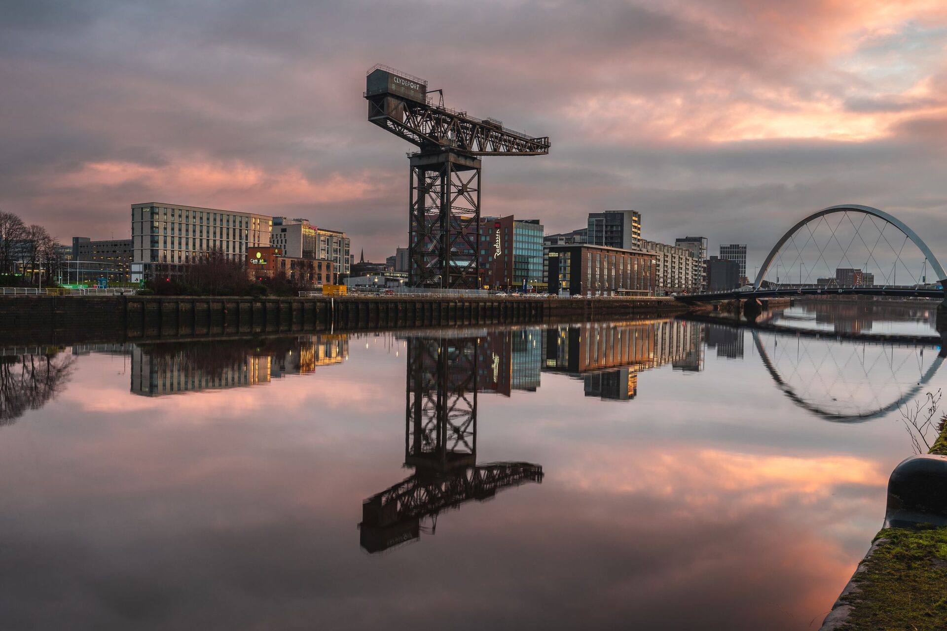 The Finnieston Crane and the arch of the Clyde Arc bridge reflected in the still River Clyde under a pink sunset sky