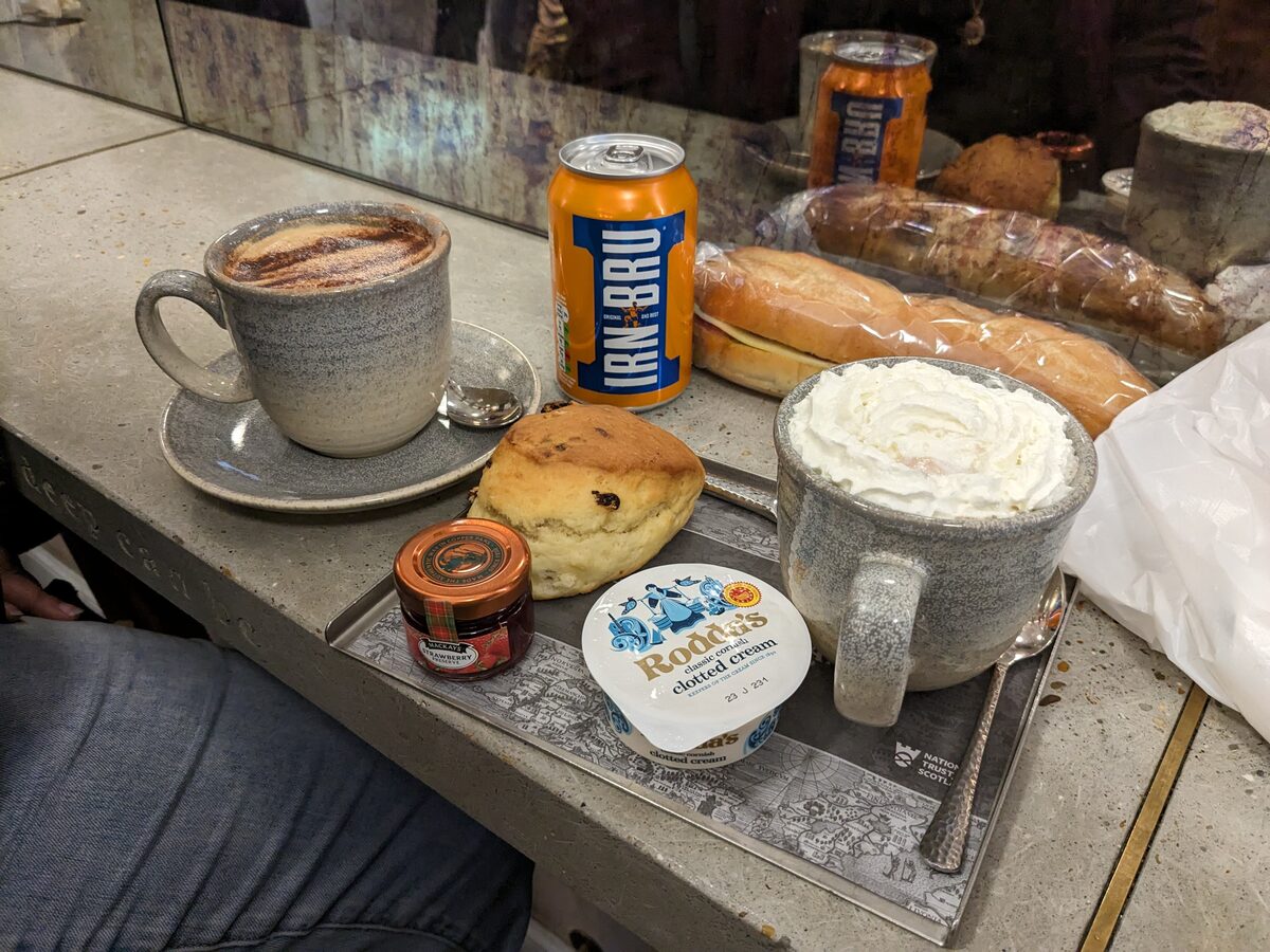 A can of Irn-Bru next to a scone with jam and clotted cream and a coffee on a café table