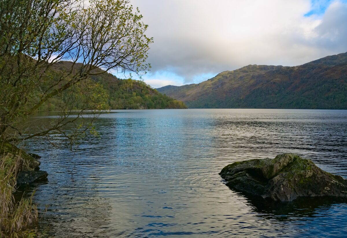 Loch Lomond on a calm day, with wooded hills along the shore and a rock in the water in front