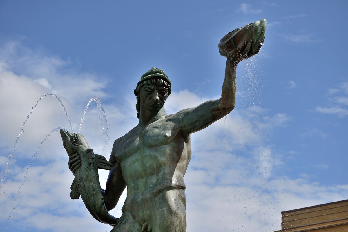 The bronze statue of Poseidon holding a fish and a shell, with water jets around him against a blue sky