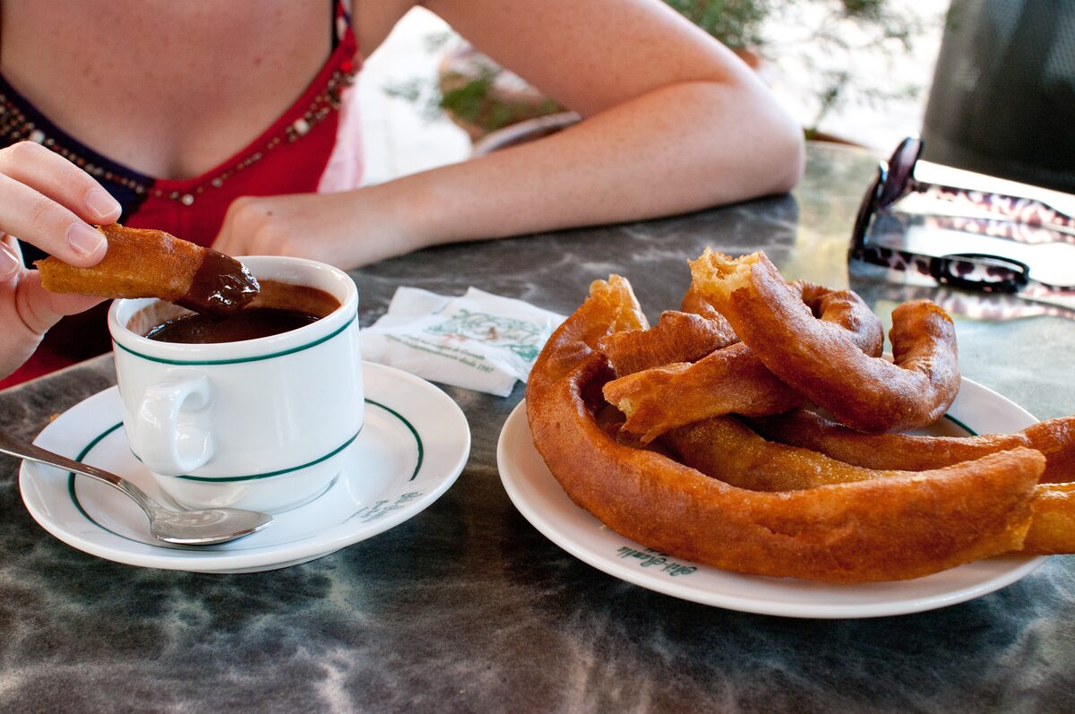 Churros with a cup of thick hot chocolate on a café table, with a hand dipping a churro