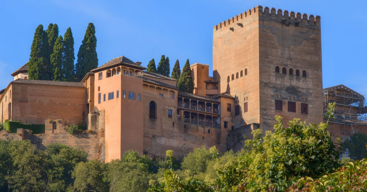 The red walls and towers of the Alhambra's Nasrid palaces rising above green trees and tall cypresses under a blue sky