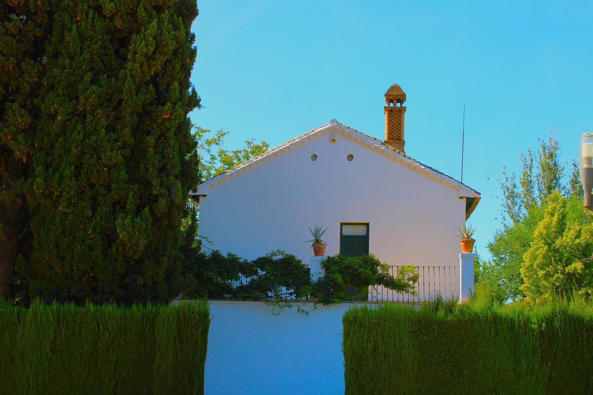 The white farmhouse of the Huerta de San Vicente with green shutters, framed by tall hedges and trees under a blue sky
