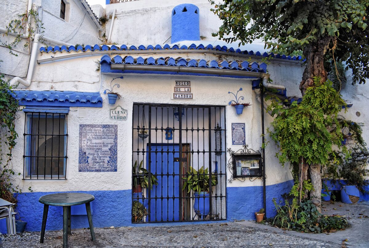 A whitewashed cave house in Sacromonte with blue doors, window frames and trim, potted plants and a tree in front