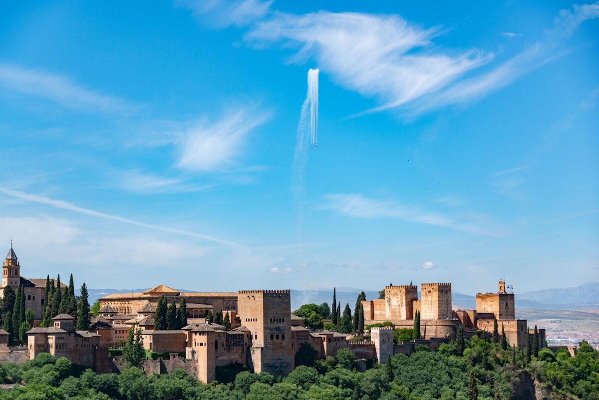 The Alhambra spread along its hill above green trees, seen from the Albaicín under a blue sky with thin clouds