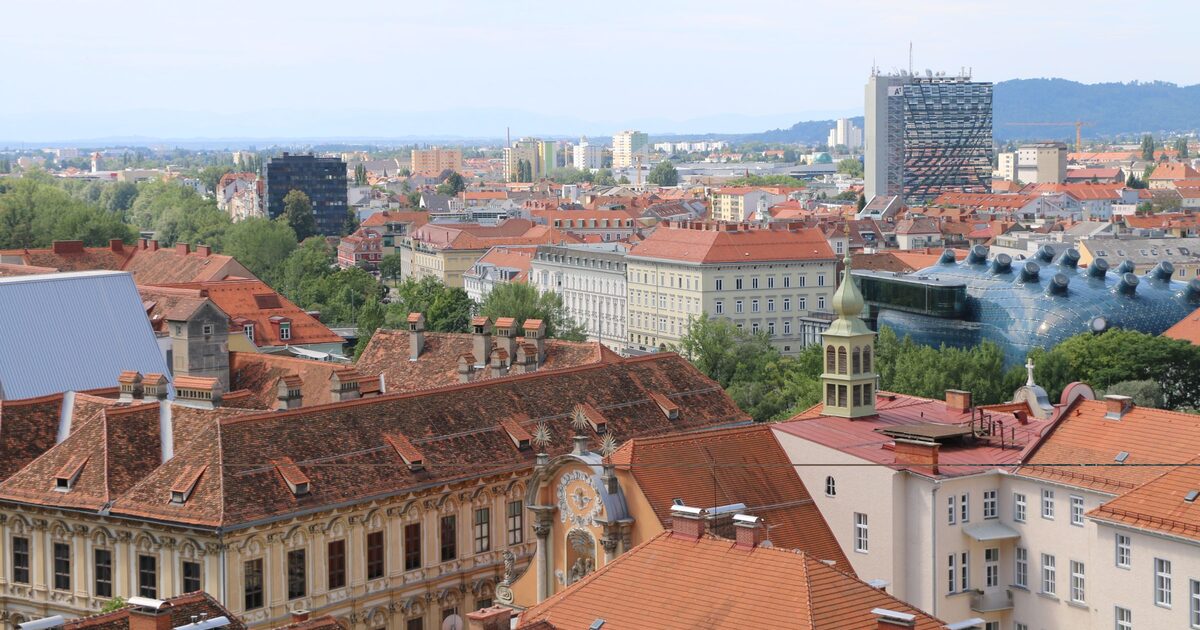 Aerial view of Graz's red-tiled old town rooftops, with the blue curved shape of the Kunsthaus by the river and modern towers behind