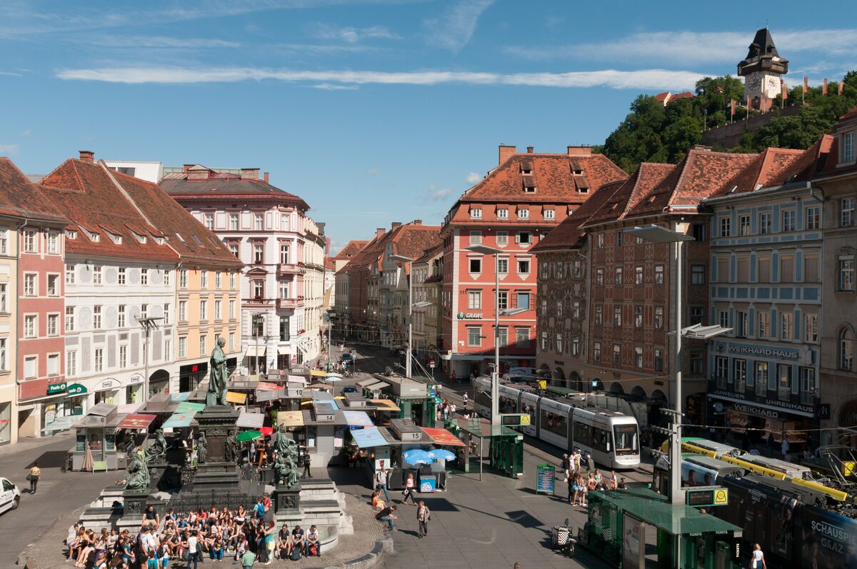 The main square of Graz seen from the town hall, with market stalls, trams, the fountain and the clock tower on the hill above