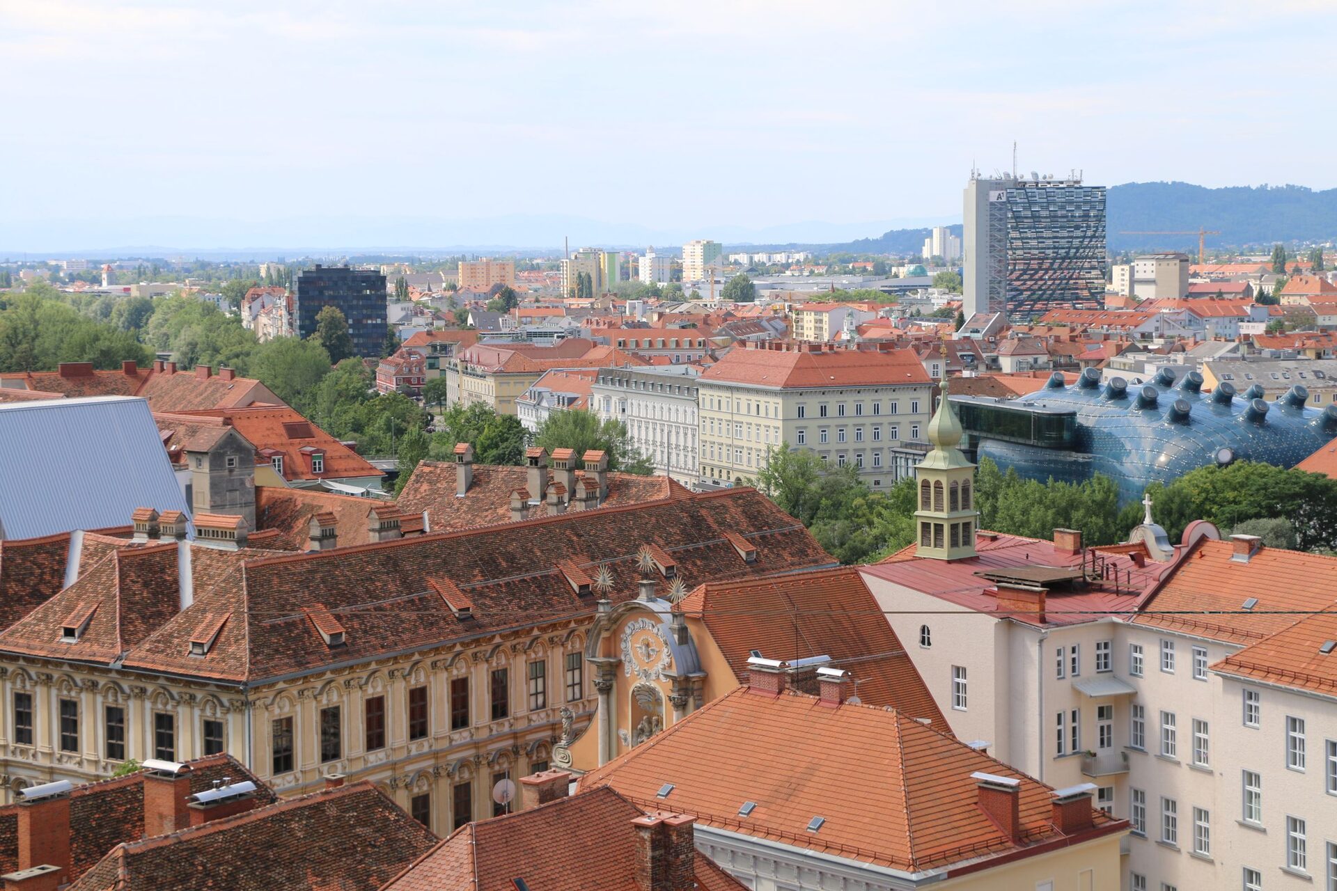 Aerial view of Graz's red-tiled old town rooftops, with the blue curved shape of the Kunsthaus by the river and modern towers behind