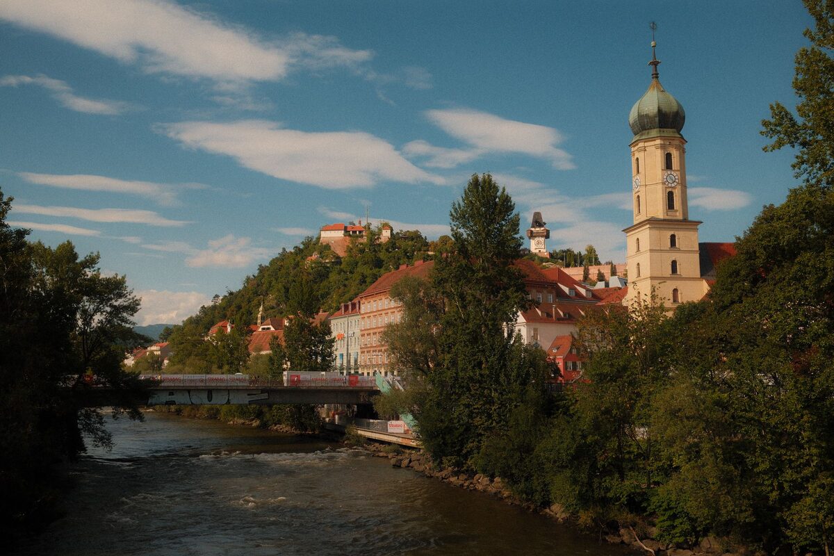 The river Mur flowing through Graz under trees, with the Schlossberg and a church tower above