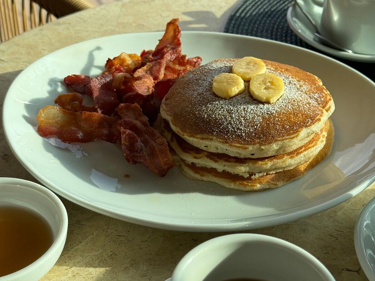 A stack of pancakes with banana and bacon beside a cup of coffee