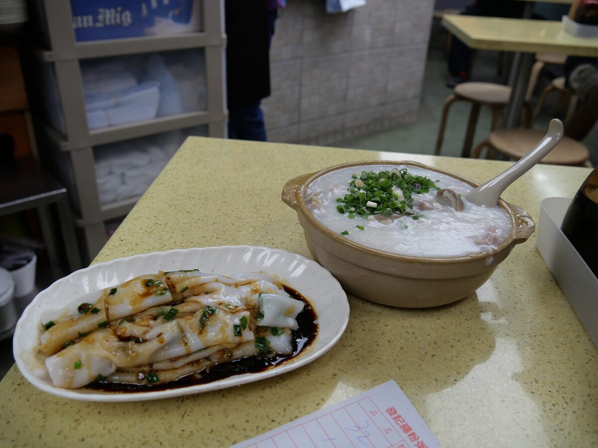 A bowl of congee with spring onion next to a plate of rice noodle rolls in soy sauce, on a cafe table
