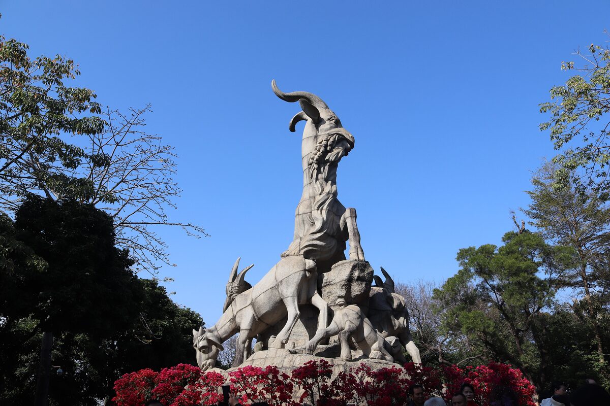 The Five Rams stone sculpture in Yuexiu Park against a blue sky, with red azalea flowers at its base