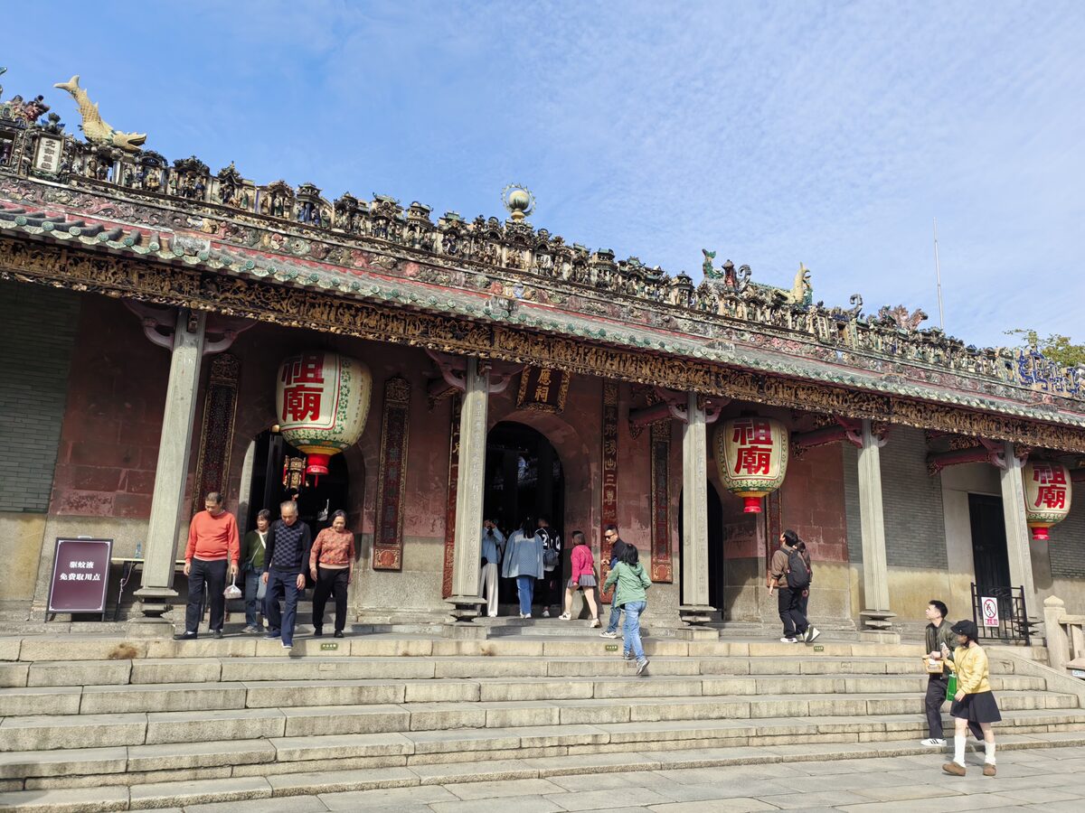 The front of the main hall at Foshan Ancestral Temple, with a carved ceramic ridge roof, red lanterns and visitors on the entrance steps