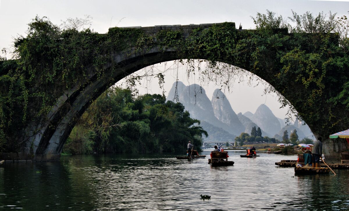 The ivy-covered stone arch of the Dragon Bridge over the Yulong River with rafts and karst peaks beyond