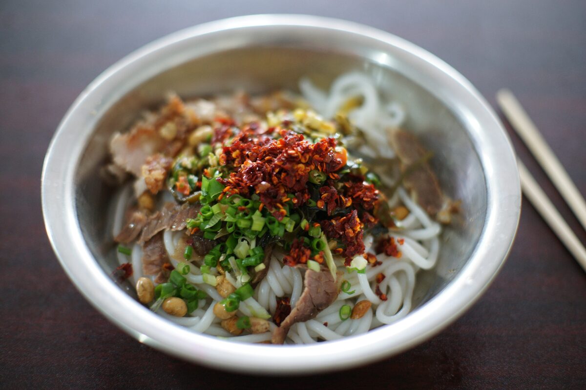 A metal bowl of Guilin rice noodles topped with sliced beef, peanuts, chopped chives and red chili
