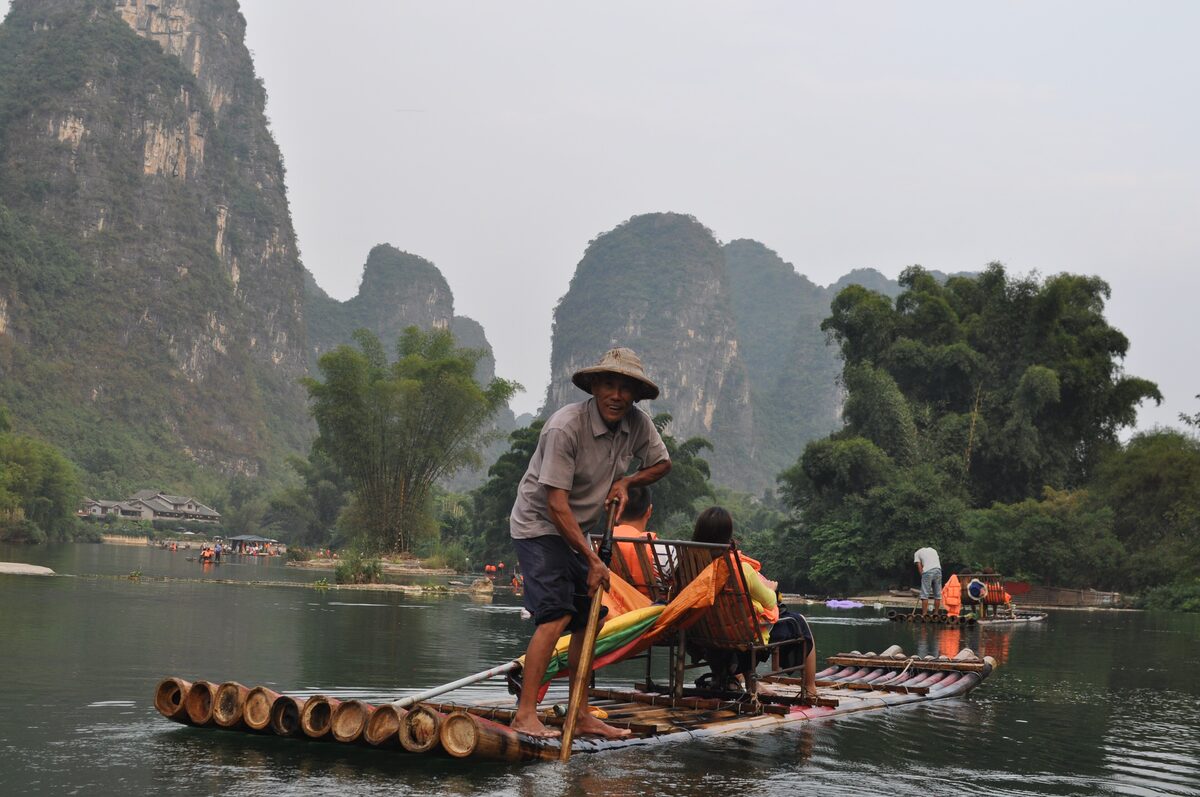 A boatman poling a bamboo raft with passengers along the Yulong River beneath karst peaks near Yangshuo