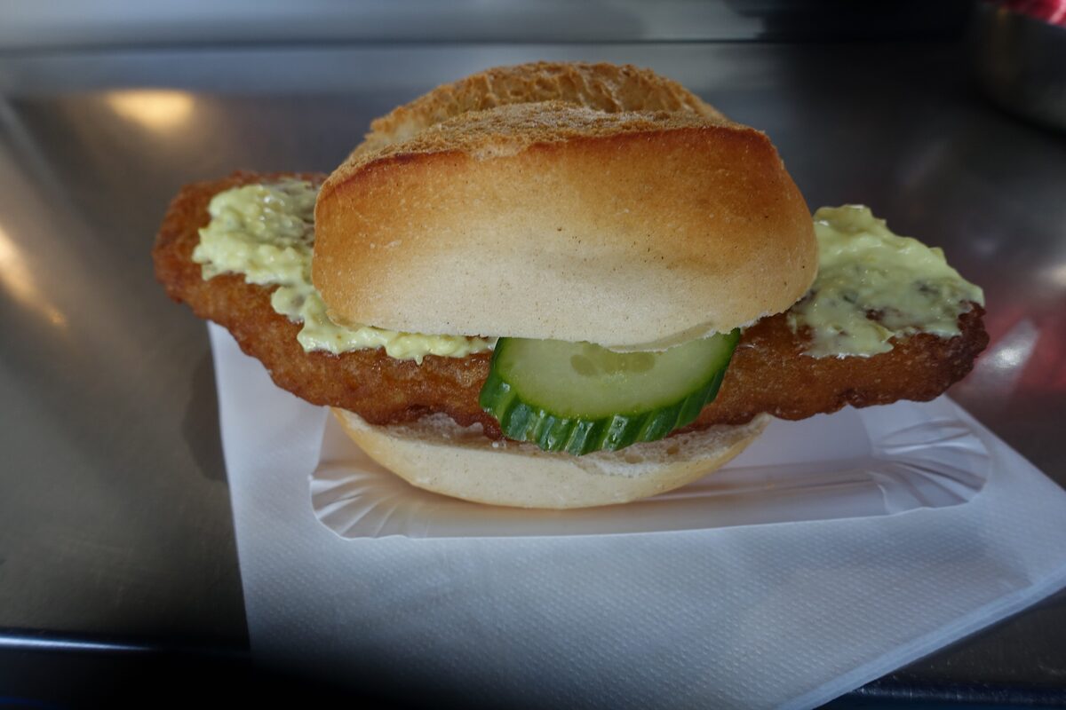 A Hamburg fish roll with fried fish, remoulade and a slice of cucumber on a paper tray