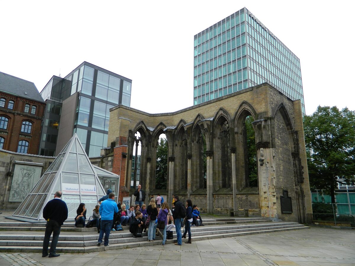 The ruined walls and arched windows of the bombed St. Nikolai church, with a glass pyramid and visitors sitting on the steps in front