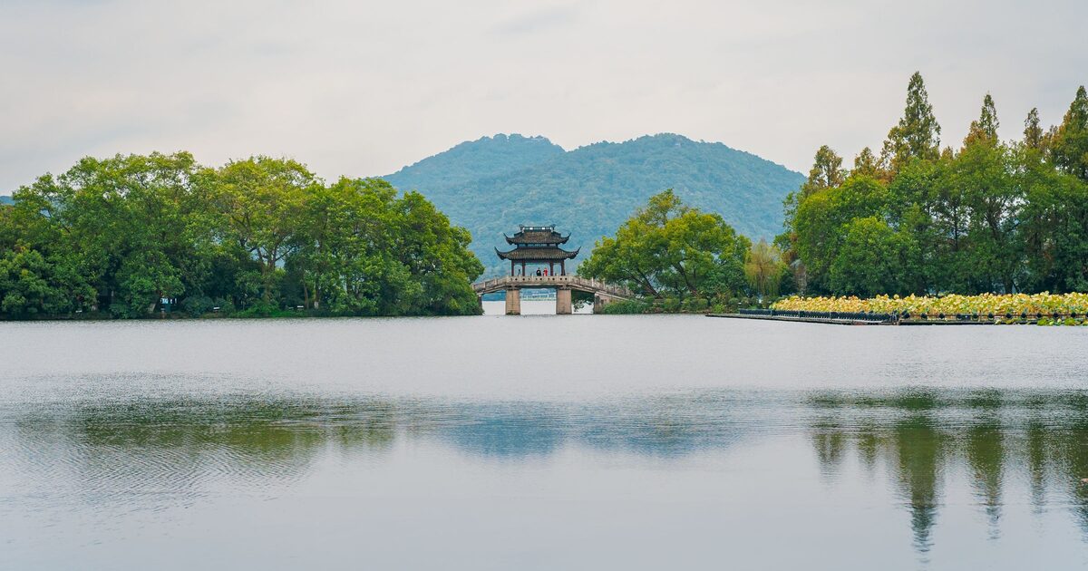 A stone causeway bridge with a two-roofed pavilion crossing West Lake, with wooded hills behind and calm water in front