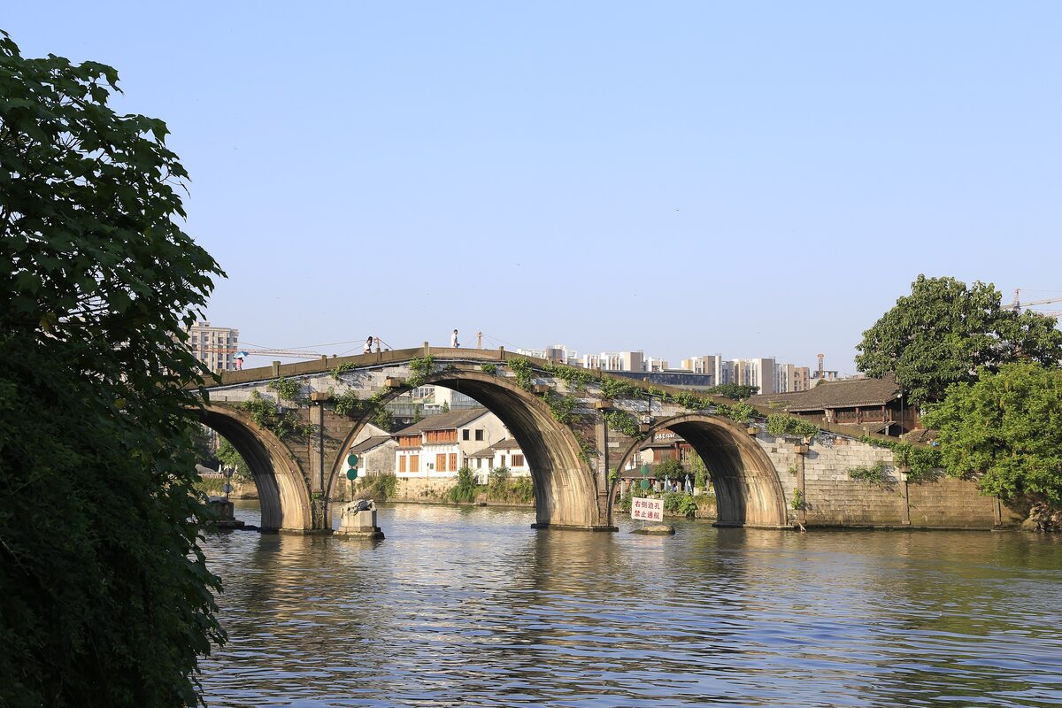 The triple-arched stone Gongchen Bridge over the Grand Canal on a clear day, with apartment blocks behind it