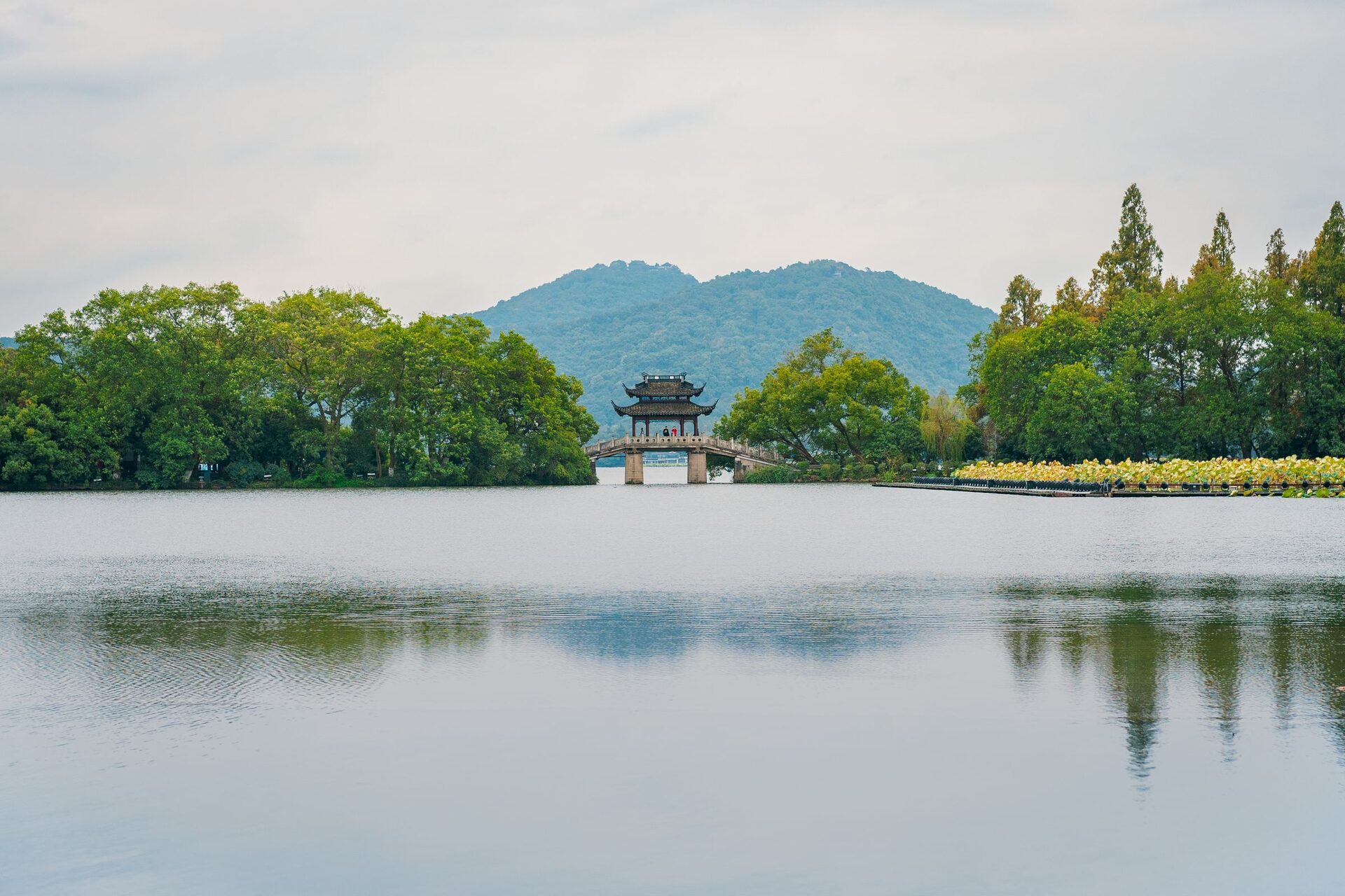 A stone causeway bridge with a two-roofed pavilion crossing West Lake, with wooded hills behind and calm water in front