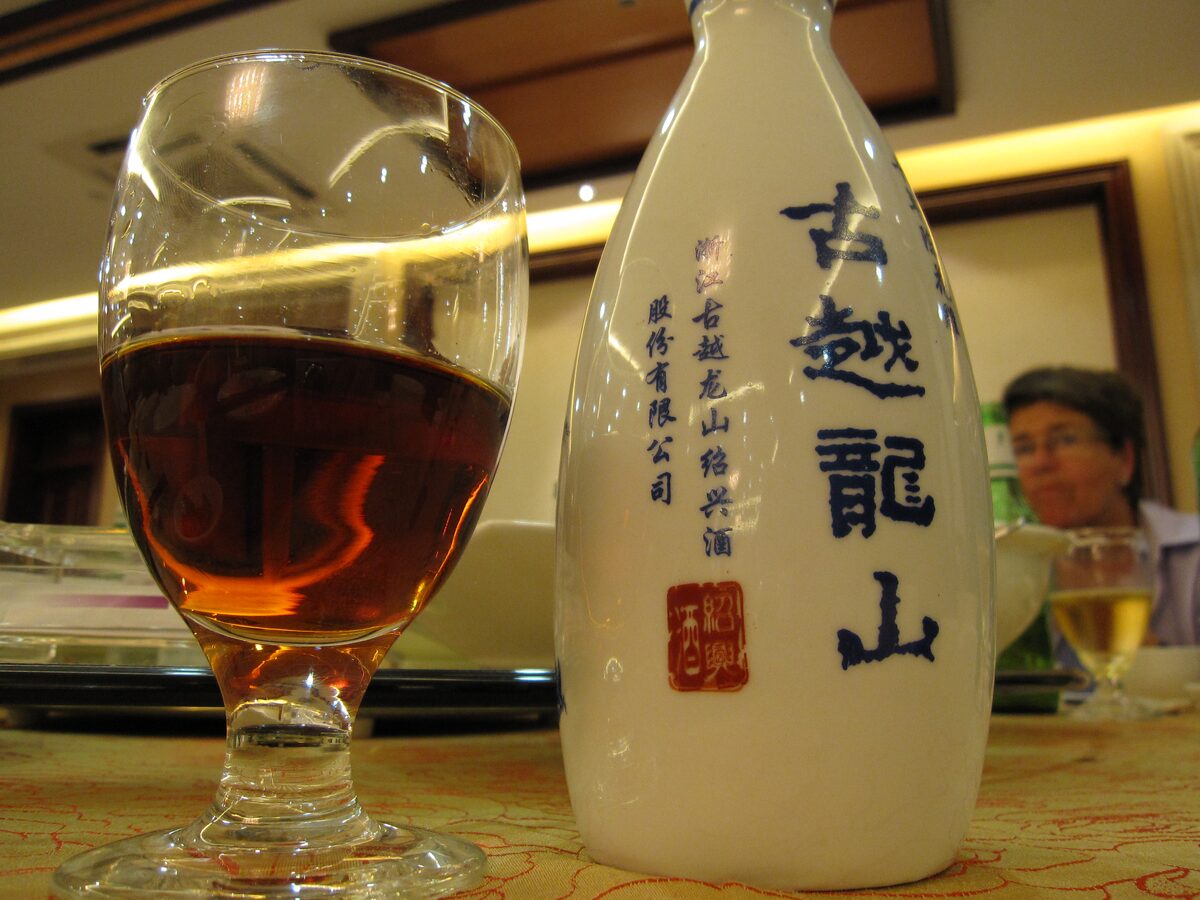 A glass of amber Shaoxing rice wine beside a white bottle on a restaurant table