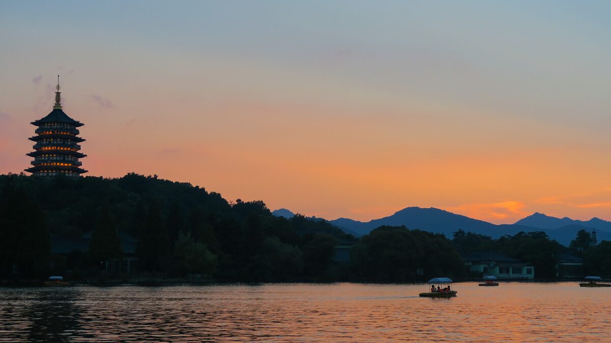Leifeng Pagoda on its hill against an orange evening sky over West Lake, with a small boat on the water