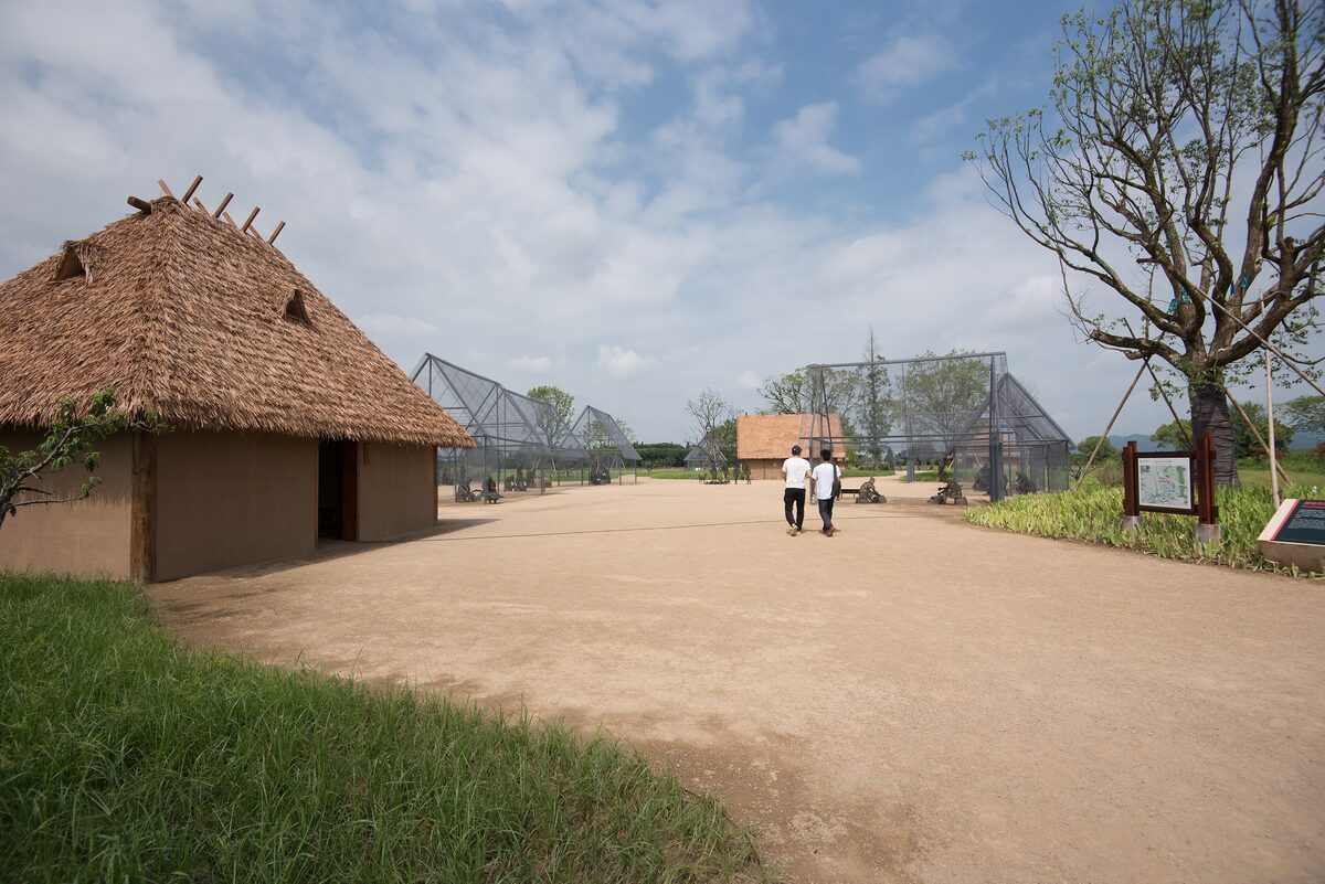 A reconstructed thatched house beside a dirt path at the Liangzhu archaeological site, with two visitors walking ahead