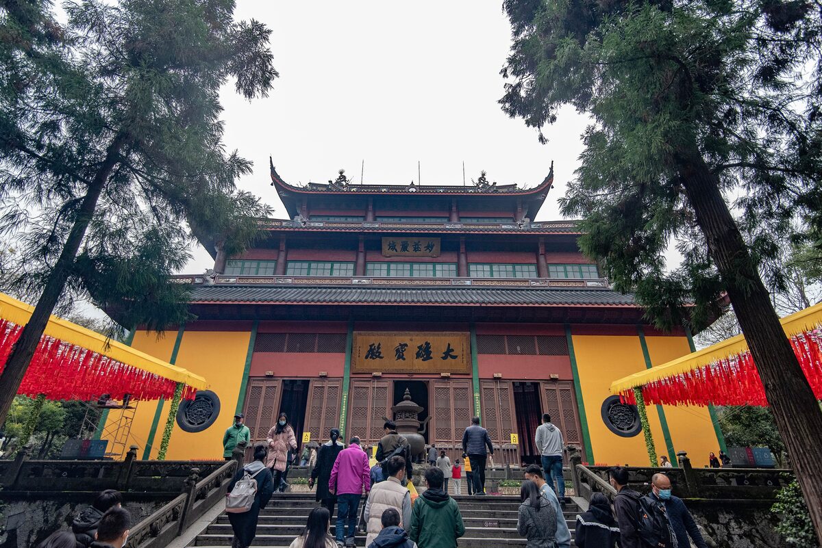 Visitors climbing the steps to the yellow and red Great Hall at Lingyin Temple, between tall trees and red banners
