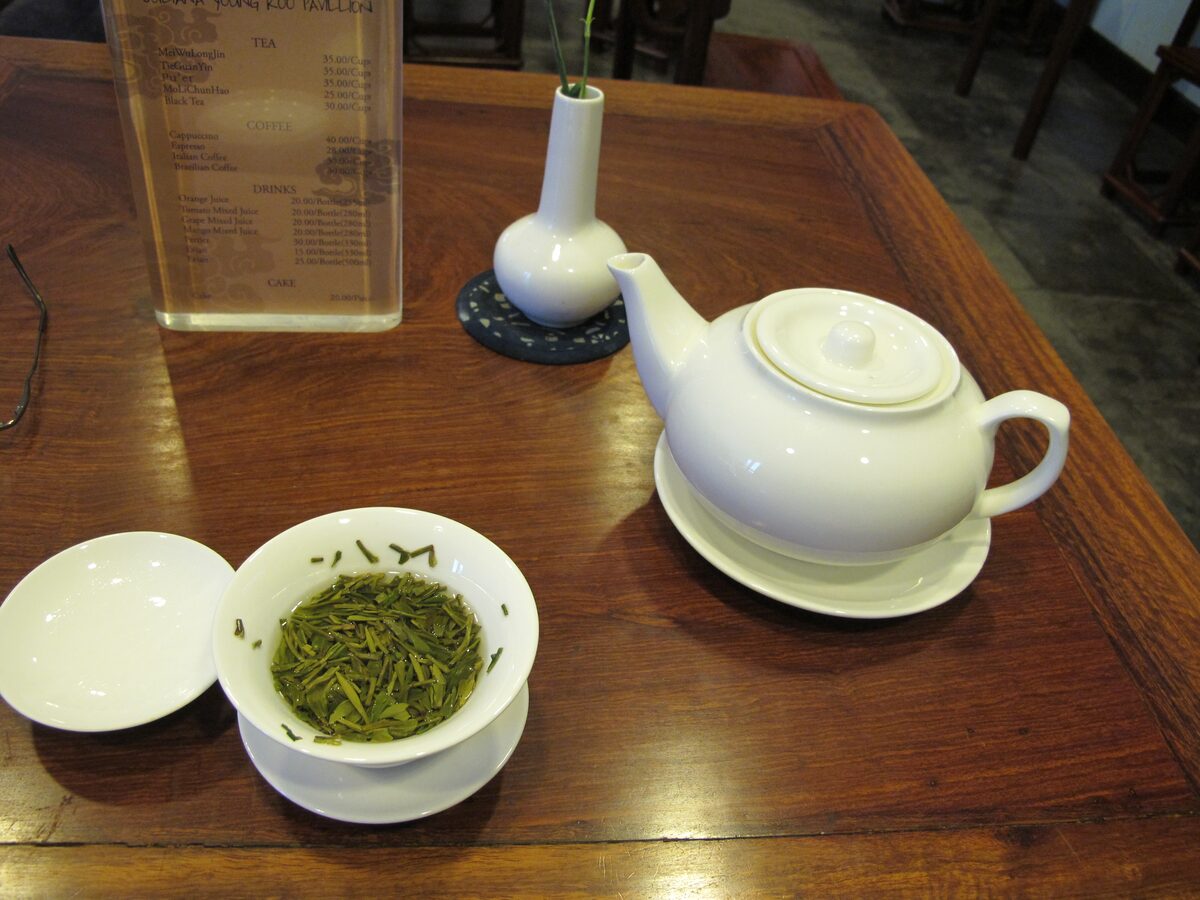 Flat green Longjing leaves in a small white cup beside a teapot on a wooden table