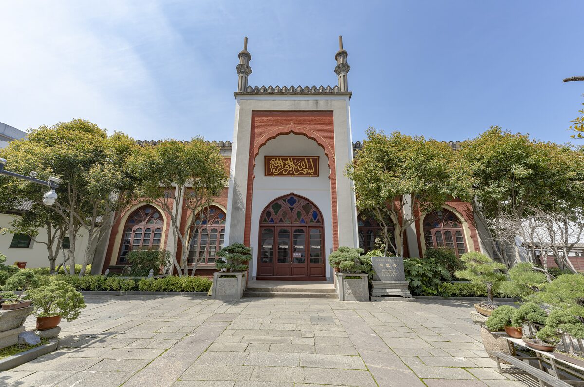 The arched entrance of the prayer hall at the Phoenix Mosque, with potted bonsai trees on the paved courtyard