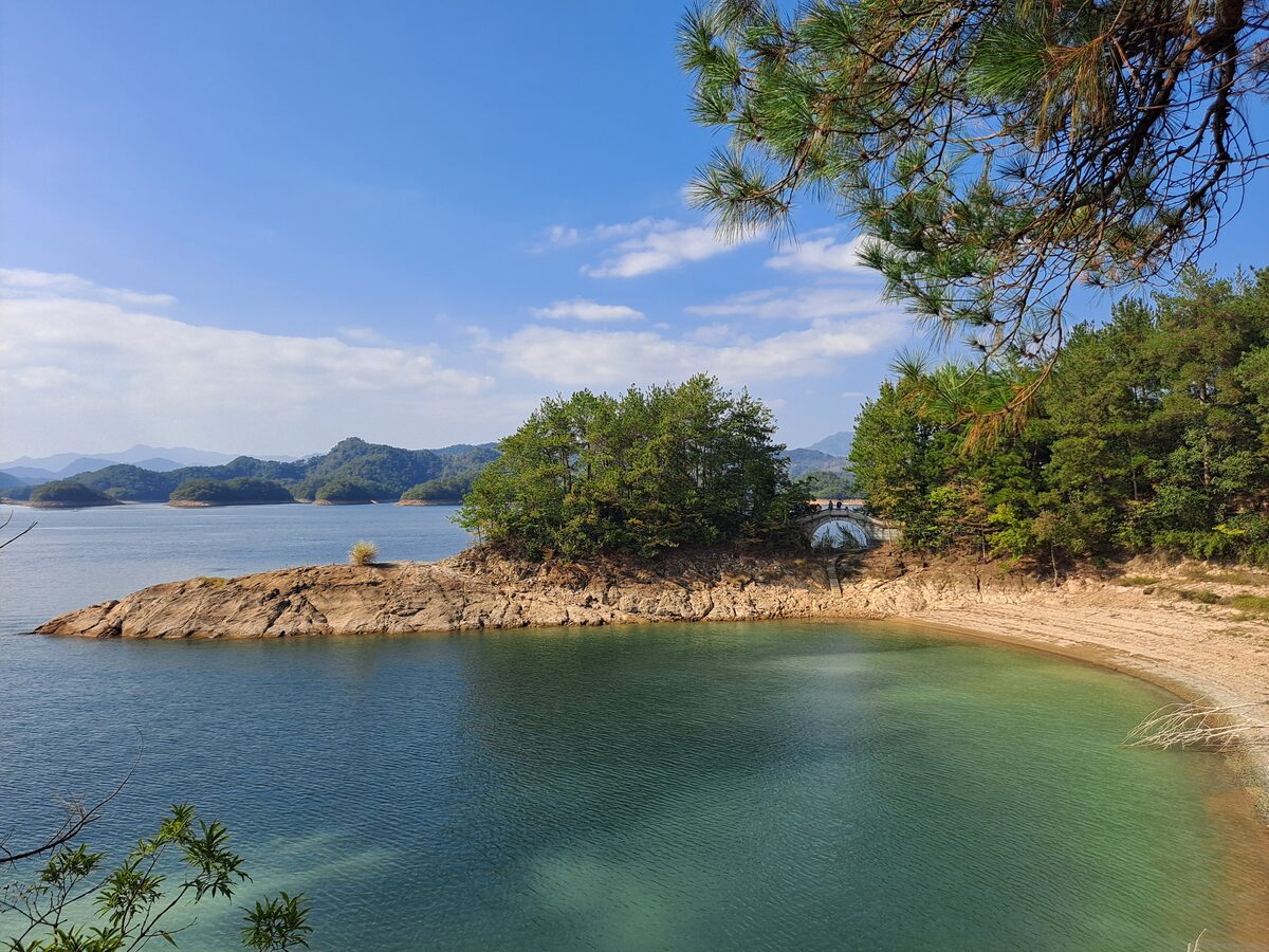 A small wooded island with a pale shore in the green water of Qiandao Lake, with more islands and hills beyond