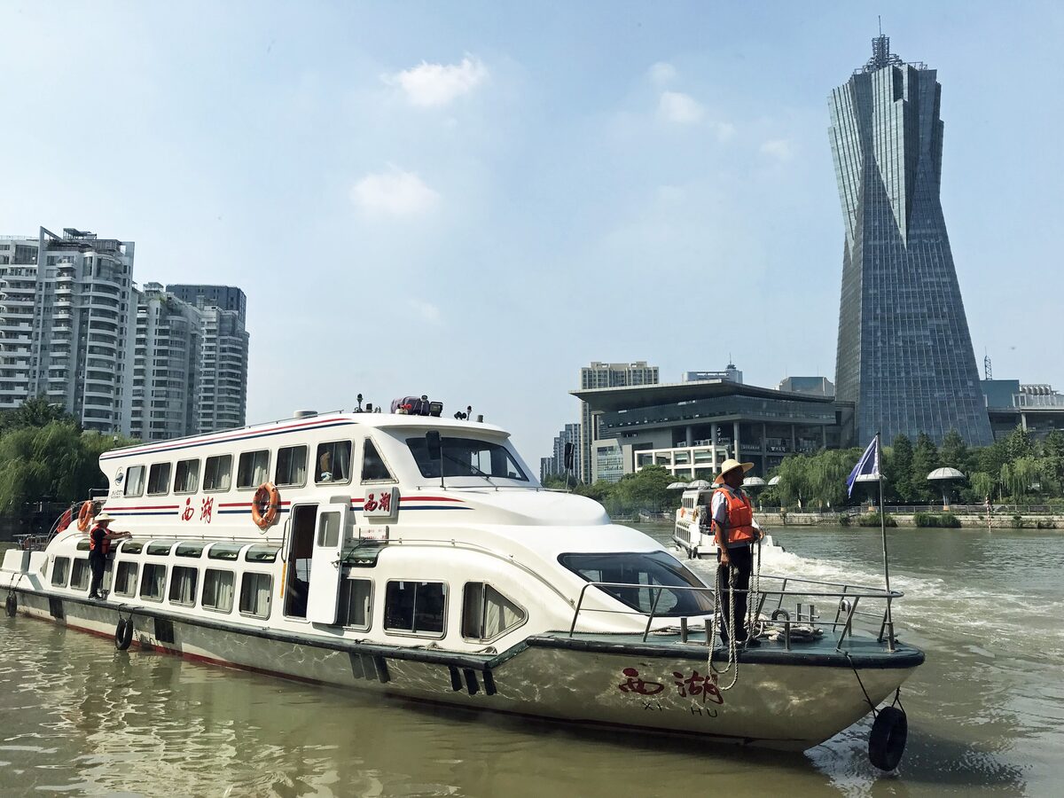 A white passenger water bus on the Grand Canal in Hangzhou with a twisted glass tower rising behind it